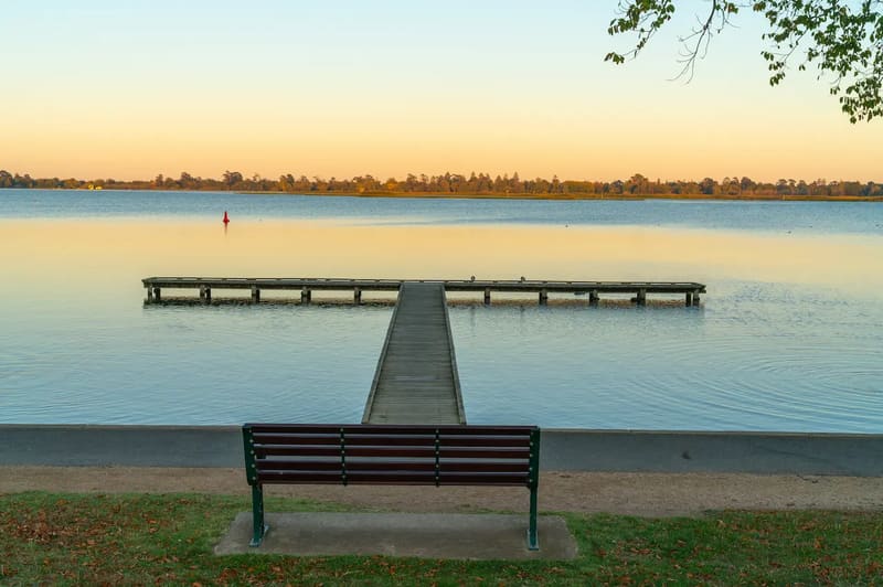 Park bench facing a timber jetty that runs out into a still lake under a golden sky